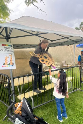 A little girl feeding a giraffe hand puppet held by an adult with a sign nearby that says, "Practice Giraffe Feeding".