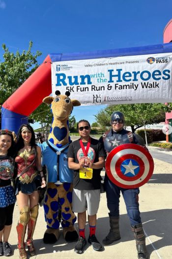An image of participants standing in front of a large arch with a sign that reads, "Run with the Heroes 5K Fun Run & Family Walk".  Of the participants standing, they are standing in a line, arm in arm, smiling at the camera.  Some are wearing mascot attire with the Valley Children's Hospital giraffe wearing a doctor's attire present and others wearing hero costumes.