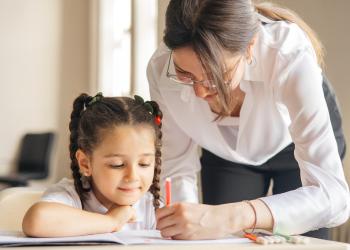 A girl sitting at a desk looking at a paper while an adult leans forward to assist the girl, providing details by writing on the girl's paper.