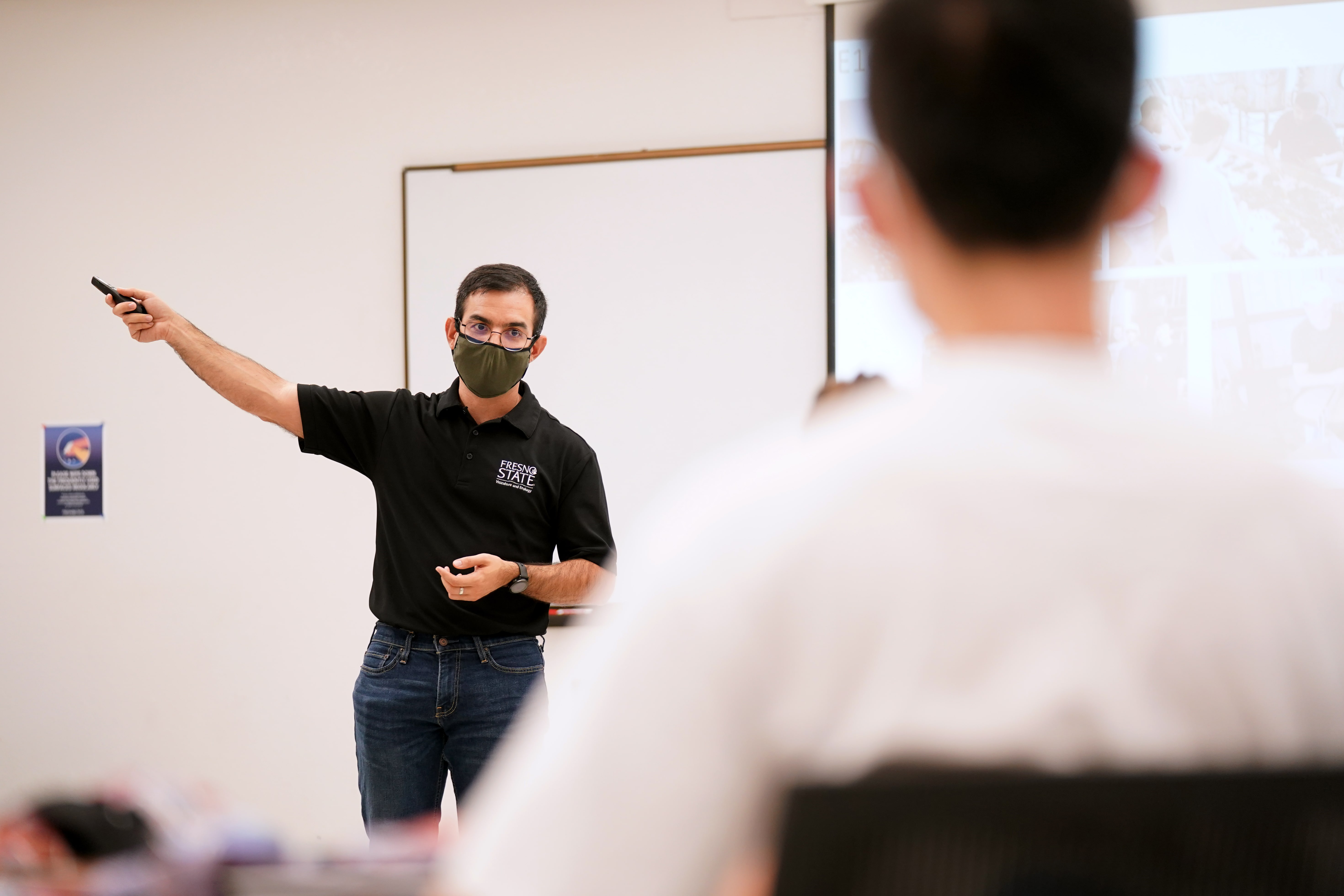 A man wearing a black polo shirt with "Fresno State" written on it and a green face mask is pointing at a screen in a classroom. He is holding a pointer or remote in his right hand. The classroom is sparsely decorated, with a whiteboard in the background. Another person, blurred, is seated in the foreground, watching the presentation.