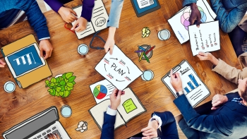 A top view of a business strategy meeting with multiple people gathered around a wooden table, hands visible. They are working with various tools such as digital tablets, smartphones, and paper documents including charts and plans.