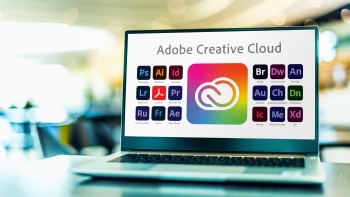 A laptop screen displaying Adobe Creative Cloud applications in a brightly lit environment. The screen shows various app icons including Photoshop, Illustrator, and Premiere Pro among others, indicating a suite of software tools used for creative projects.